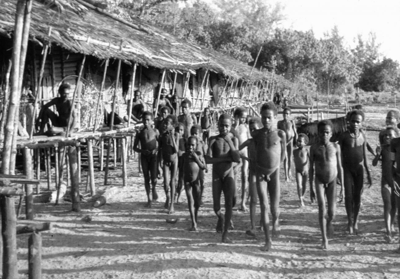 BD/216/161 -
Groep lopende kinderen, gadegeslagen door enige ouderen, zittend op de waranda