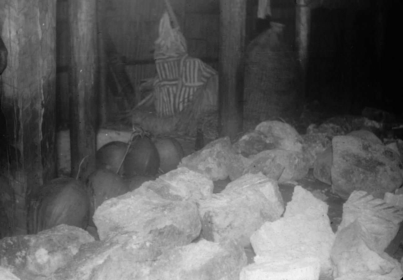BD/216/92 -
Interieur huis met op voorgrond sago en kokosnoten en achtergrond 2 Jipae kostuum dansmaskers
