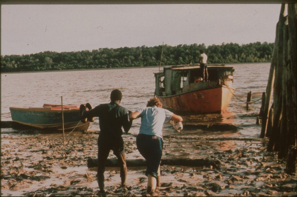 BD/30/64 - 
Asmat man helps white woman crossing the mud to a boat on the river
