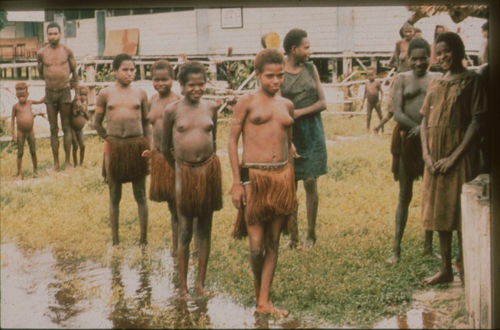 BD/30/96 - 
Asmat girls in bark cloth skirts standing in the marsh in front of a large building
