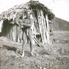 BD/329/44 Papoea-man poseert voor een hut met een varkentje in zijn hand